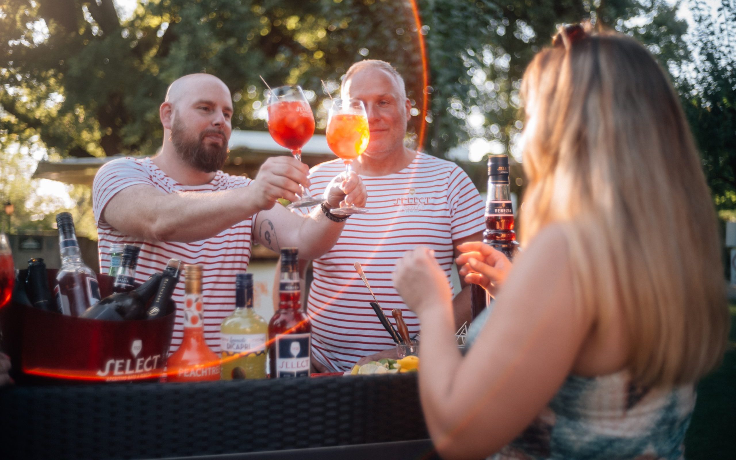 Zwei Männer in gestreiften T-Shirts überreichen Aperitif-Gläser an eine Frau mit langen Haaren, die ihnen gegenüber an einer Outdoor-Bar steht. Im Vordergrund sind verschiedene Flaschen von Spirituosen und Likören zu sehen. Im Hintergrund sind Bäume und Sonnenlicht zu erkennen, was eine entspannte Sommeratmosphäre schafft.