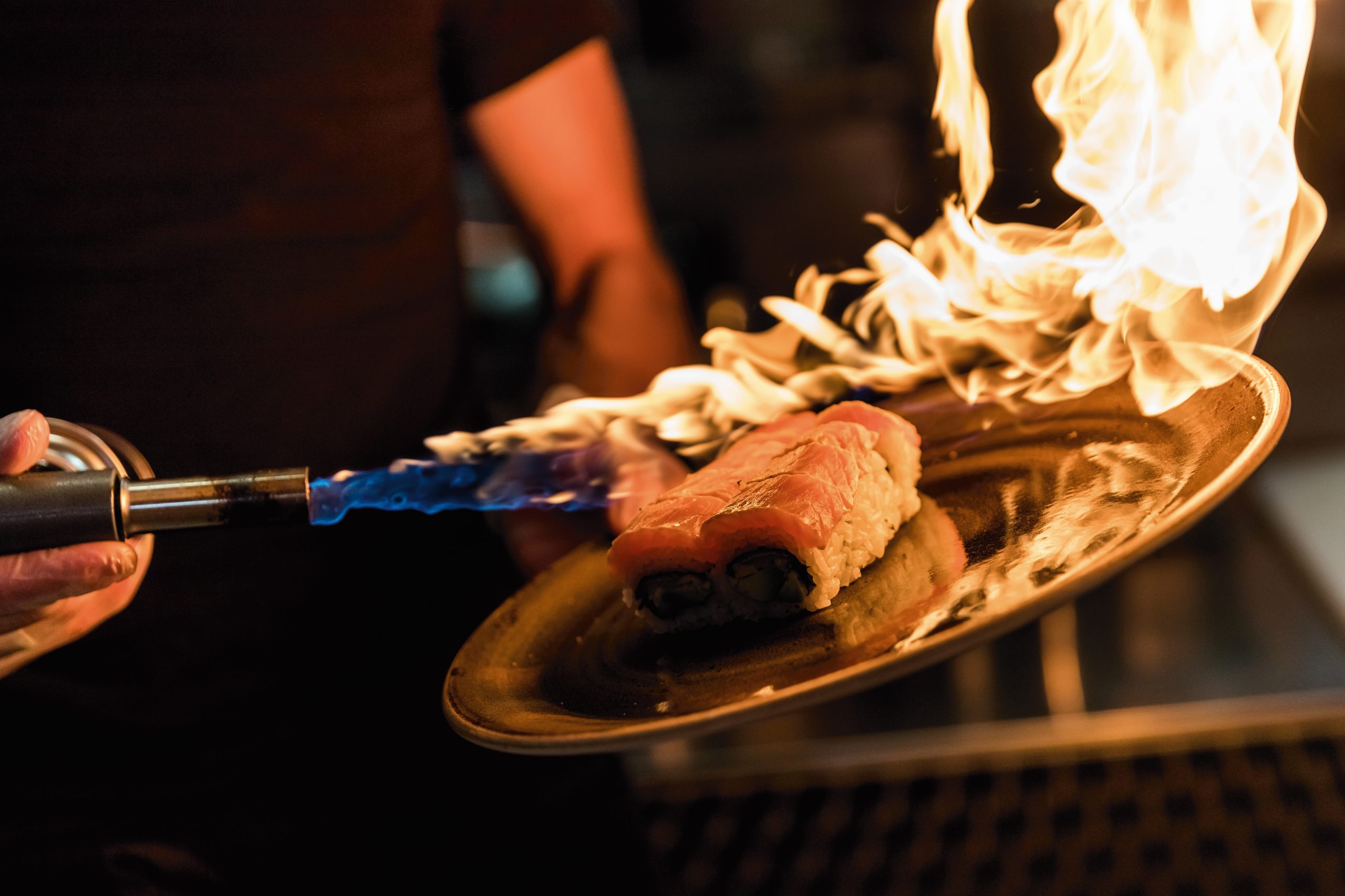 Ein Koch flambiert ein Sushi-Gericht mit einem kleinen Flammenwerfer in einer Küche.