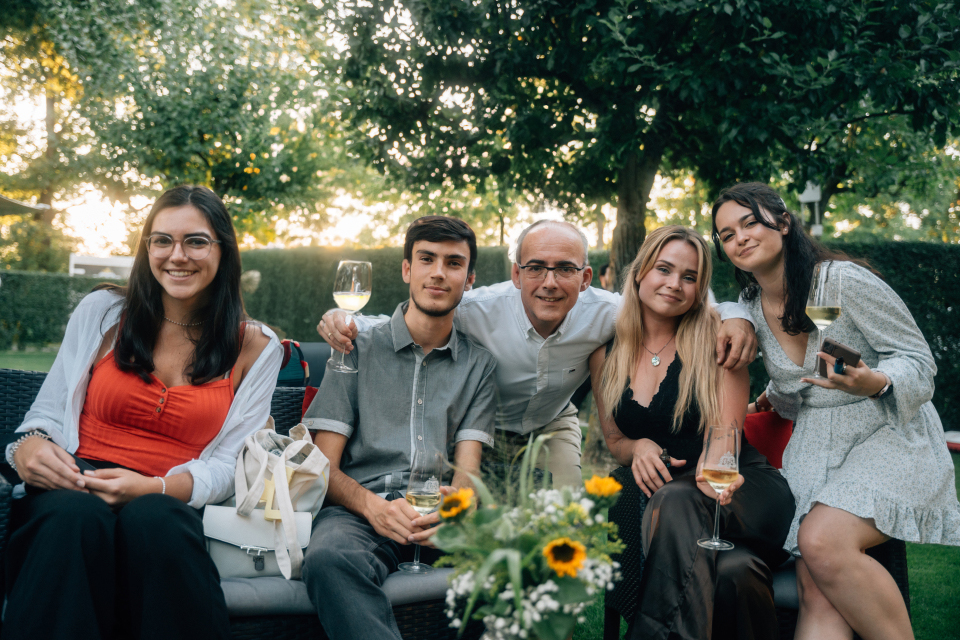 Eine Gruppe von fünf Personen sitzt draußen auf einer Gartenbank. Sie lächeln und halten Weingläser in den Händen. Im Hintergrund sind Bäume und Sträucher zu sehen, die eine entspannte Atmosphäre im Freien schaffen.