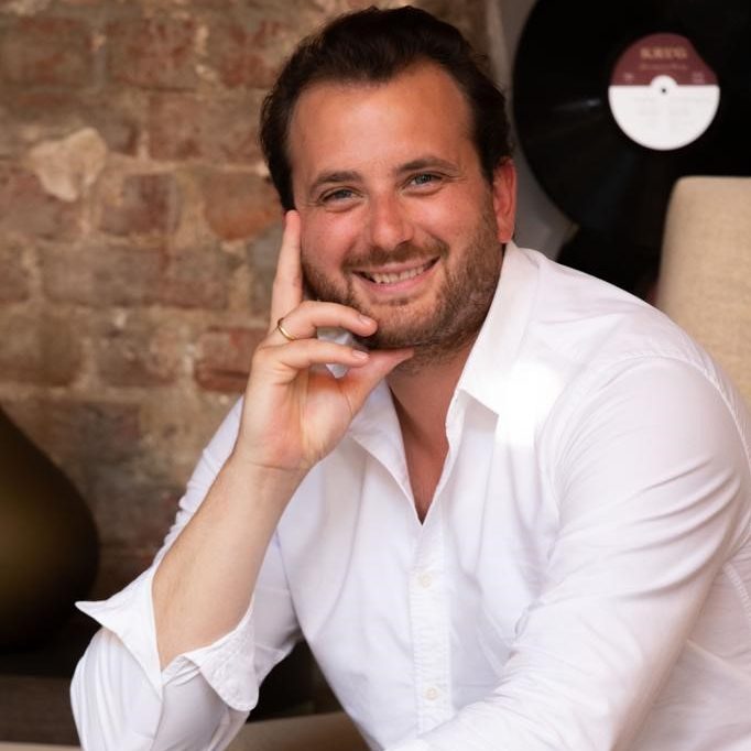 Ein Mitarbeiter sitzt lächelnd in einem gemütlichen Raum. Er trägt ein weißes Hemd und eine Uhr am Handgelenk, seine Hand stützt sein Kinn, während er in die Kamera blickt. Im Hintergrund ist eine Ziegelwand und ein Vinyl-Schallplattenständer zu sehen.