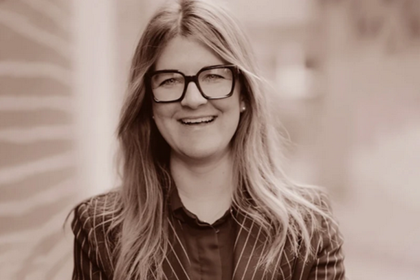 Brand Managerin Fine Wine Eine Mitarbeiterin mit mittellangem, blonden Haar und Brille lächelt in die Kamera. Sie trägt einen dunklen Blazer und steht vor einem unscharfen Hintergrund.