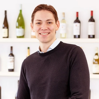 Ein Mitarbeiter lächelt freundlich in die Kamera. Im Hintergrund sind Weinflaschen zu sehen.