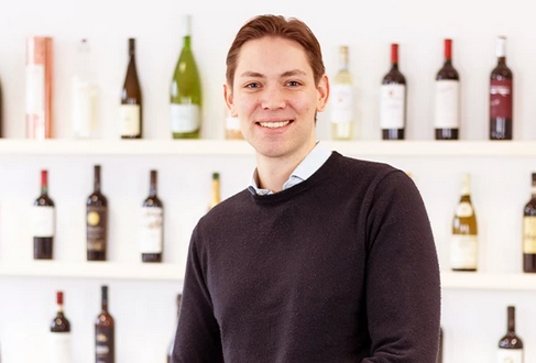 Category Manager, Marketingkoordination & Datenmanagement Ein Mitarbeiter lächelt freundlich in die Kamera. Im Hintergrund sind Weinflaschen zu sehen.