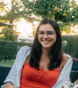 Auszubildene im Garten Eine junge Frau mit dunklem, schulterlangem Haar und Brille lächelt in die Kamera. Sie trägt ein rotes Top mit Knöpfen und eine weiße Jacke, sitzt draußen auf einem Gartenstuhl und genießt den sonnigen Tag. Im Hintergrund sind Bäume und eine Hecke zu sehen, was eine entspannte und freundliche Atmosphäre schafft.