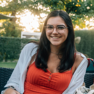Eine junge Frau mit dunklem, schulterlangem Haar und Brille lächelt in die Kamera. Sie trägt ein rotes Top mit Knöpfen und eine weiße Jacke, sitzt draußen auf einem Gartenstuhl und genießt den sonnigen Tag. Im Hintergrund sind Bäume und eine Hecke zu sehen, was eine entspannte und freundliche Atmosphäre schafft.