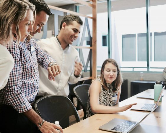 Vier Kollegen stehen um einen Schreibtisch und arbeiten gemeinsam an einem Projekt. Sie schauen konzentriert auf den Bildschirm eines Laptops und diskutieren angeregt.